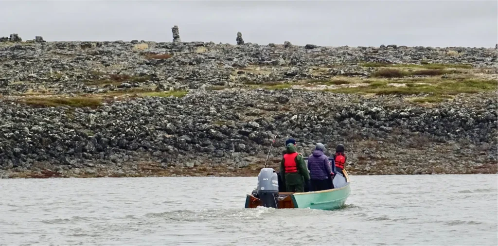 A small group of people in a motorized boat on a calm body of water, heading towards a rocky shoreline. The passengers are dressed warmly and wearing red life jackets, with inukshuks visible in the background on the rugged terrain