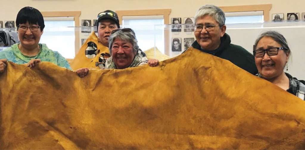 A group of five smiling individuals from Kugluktuk proudly holding up a large piece of tanned moose hide inside a community space
