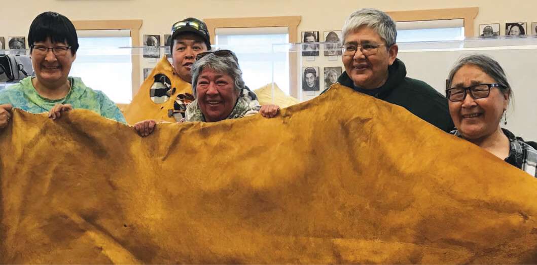A group of five smiling individuals from Kugluktuk proudly holding up a large piece of tanned moose hide inside a community space