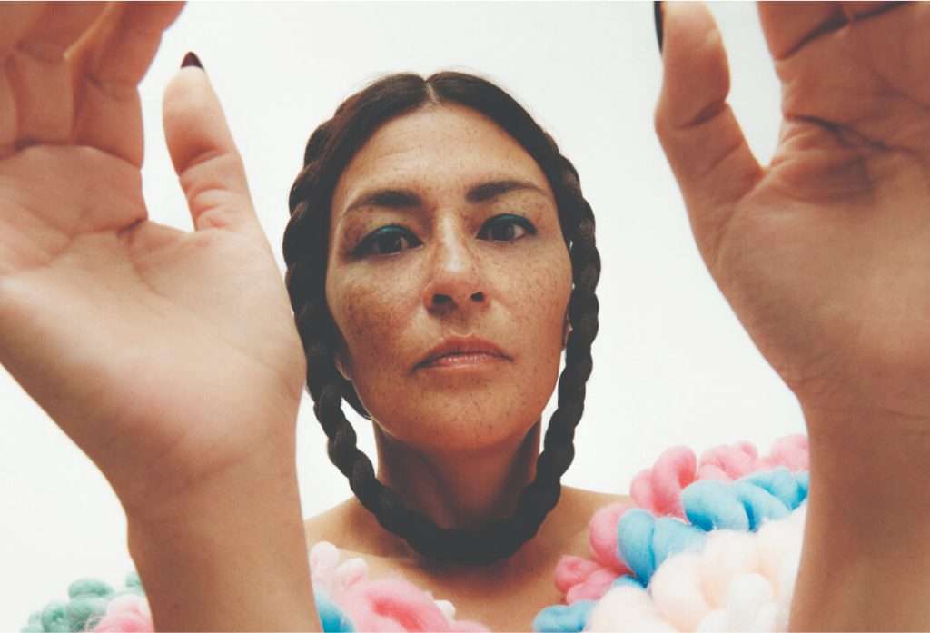 A close-up image of Elisapie, a woman with braided hair, looking directly into the camera. Her hands are raised, framing her face. She wears a colorful outfit made of pink, blue, and white elements, adding an artistic and vibrant touch to the photo. The image conveys a sense of strength, culture, and individuality.