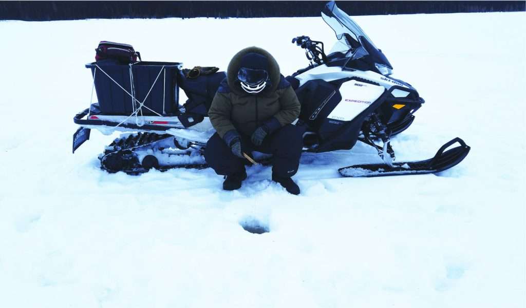 A person dressed in winter gear kneeling beside a snowmobile while ice fishing.