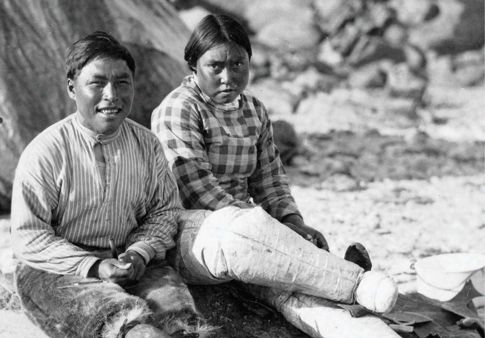Black and white photo of an Inuit couple sitting outdoors, dressed in traditional clothing
