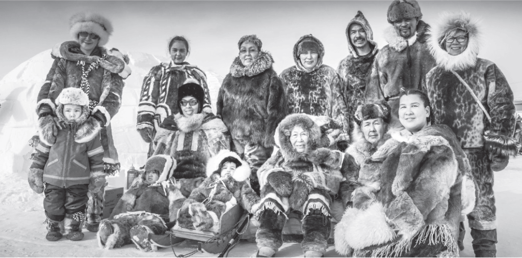 A black-and-white group photo of family members dressed in traditional Inuit clothing. The group includes adults and children, standing and sitting in front of an igloo, with some seated on a sled. Each person is warmly dressed in fur clothing, smiling for the camera.