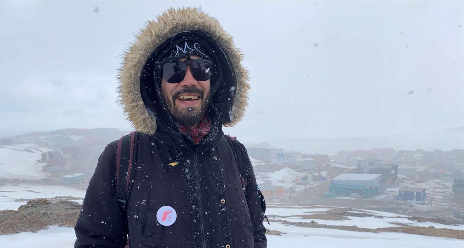 Jamesie Fournier smiling outdoors in a snowy landscape, wearing a black winter coat with a fur-lined hood, sunglasses, and a beanie. Snow is falling, and buildings are visible in the background, partially covered by snow