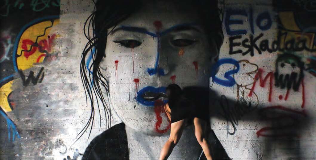 A woman appears to interact with a large, graffiti-style mural of a face on a concrete wall. The mural features bold colors and abstract lines, while the woman, dressed in black, bends forward, blending into the art.