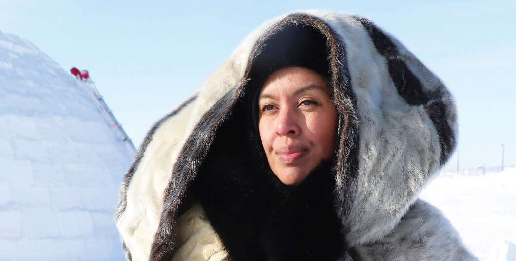 A woman dressed in traditional Inuit fur clothing, standing outdoors with an igloo in the background. She gazes into the distance under the bright, clear sky.