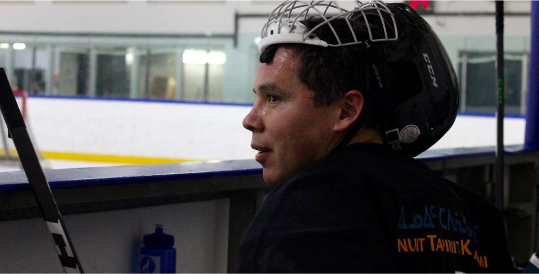 A hockey player wearing a black helmet and gear takes a break on the bench, looking out over the ice rink. The player, slightly flushed from exertion, rests between shifts during a game. The rink in the background is brightly lit, with a clear view of the ice and surrounding glass panels. The player seems focused, reflecting the intensity and passion for the game.