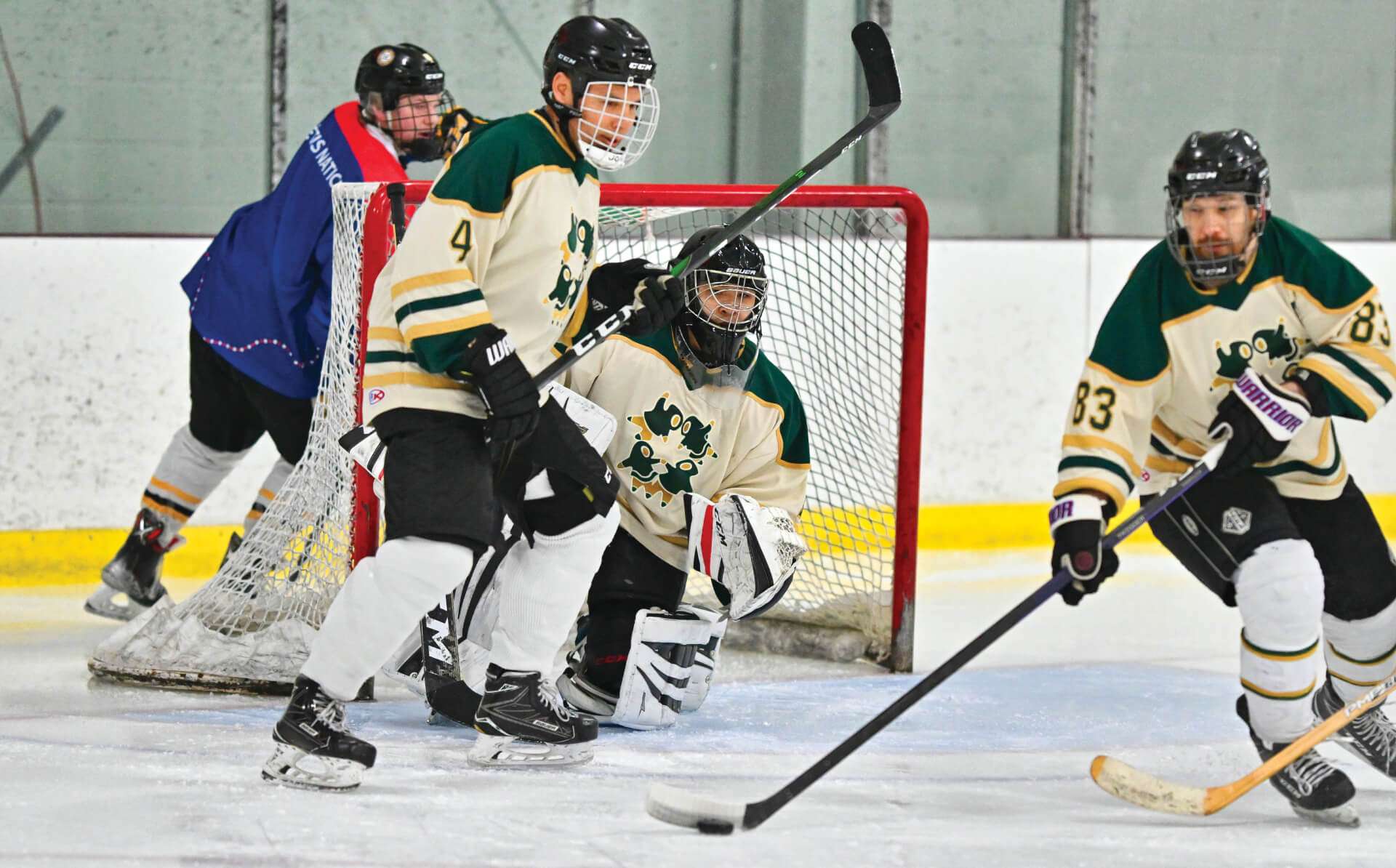 Hockey players are in action on the ice near the goalpost. A goalie in black and white gear crouches in front of the net, ready to block a shot. Two players in green and cream jerseys position themselves defensively while another player in a purple jersey skates behind the net. The intensity of the game is evident as the players focus on the puck.