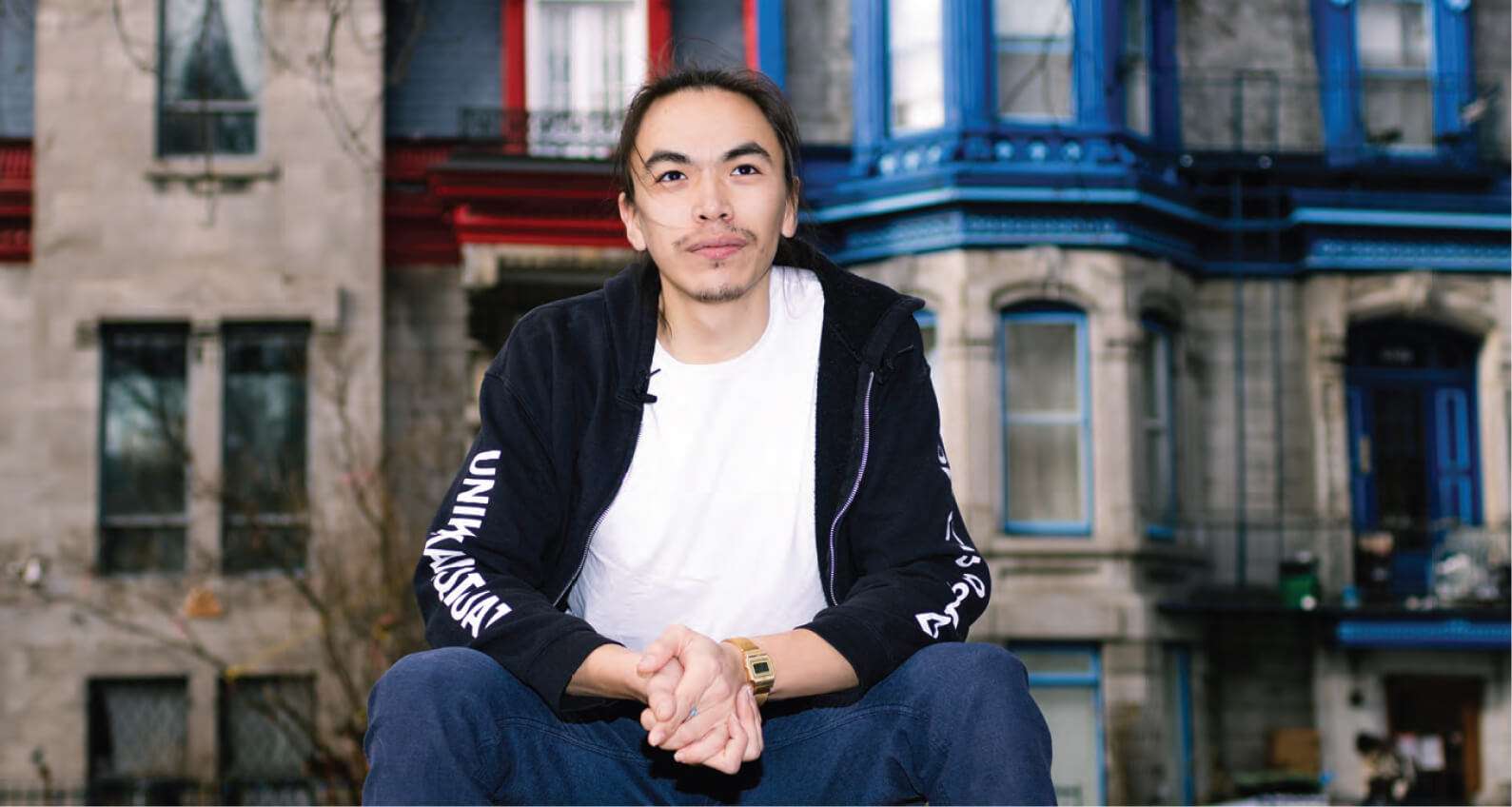 Saali Kuata, a Montreal-based Inuit multidisciplinary artist, sitting casually in front of a colorful Montreal building. He is wearing a black hoodie and a white t-shirt, looking confidently into the camera, with long hair tied back. The background features a typical Montreal street scene with stone facades and blue and red architectural accents.