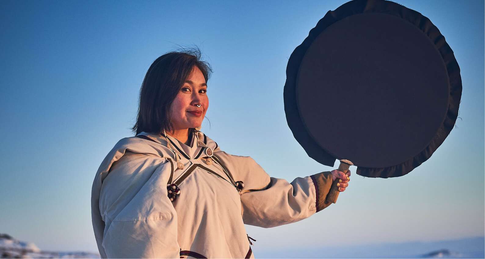 Sandi Vincent holding a traditional Inuit drum against a clear sky, wearing a light-colored parka, during a performance in Iqaluit.