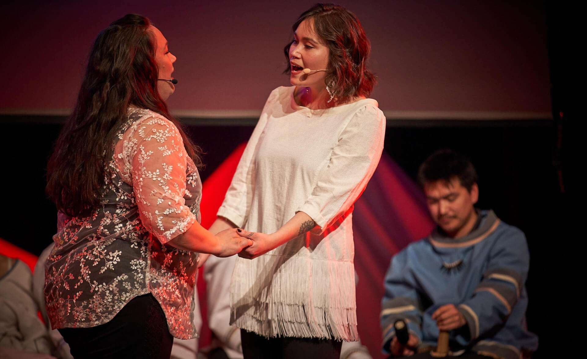 Sandi Vincent, in a white fringed outfit, performing a duet on stage with a fellow performer wearing a floral top. Both are holding hands and wearing headset microphones. Another performer is seated in the background, wearing traditional clothing.